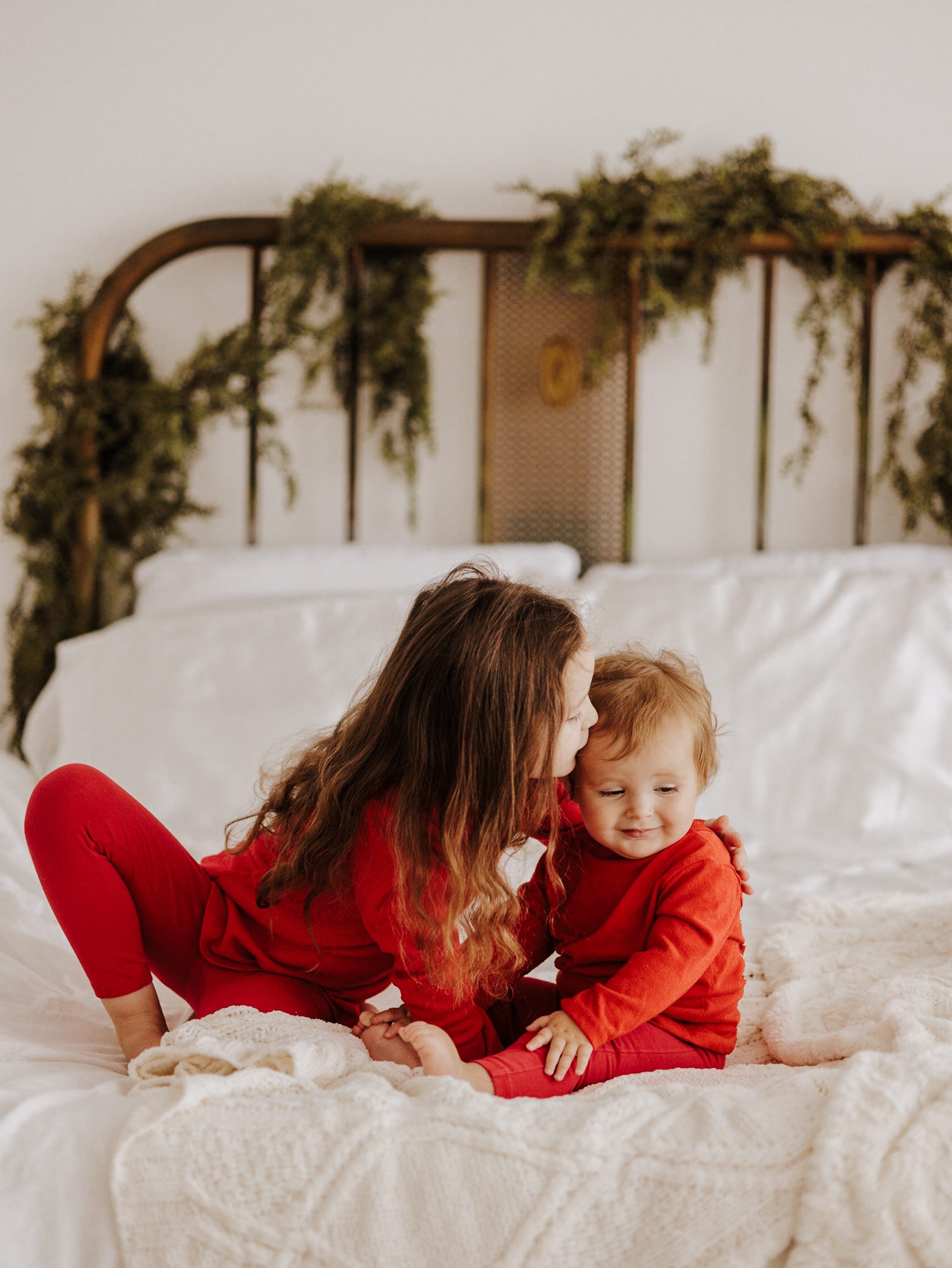 Big sister and little sister sitting on a vintage bed covered in greenery and a white bedspread. Holiday PJ Mini Sessions in Murfreesboro TN