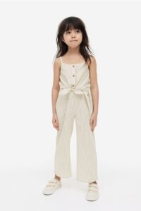 Product photo of a little girl with dark hair wearing a cream jumpsuit that ties in the front and has button details