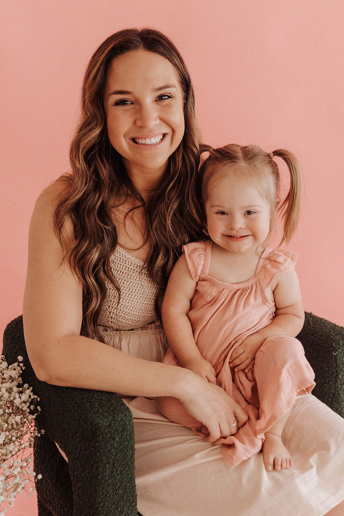 Mother and Daughter sitting in front of a pink backdrop in a green chair next to flowers for a Mother's Day Mini Session
