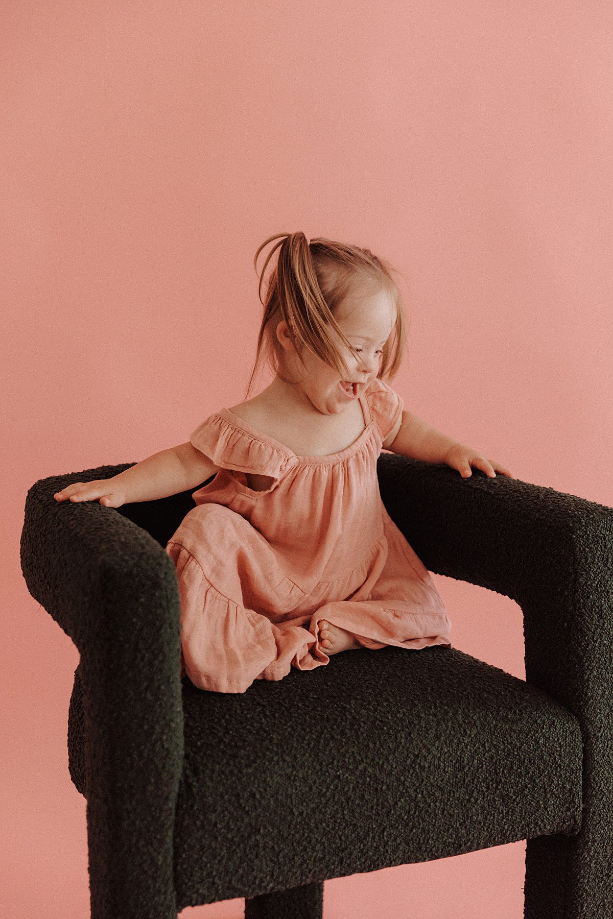 Toddler girl with Down syndrome laughs while sitting in a green chair during a mini session