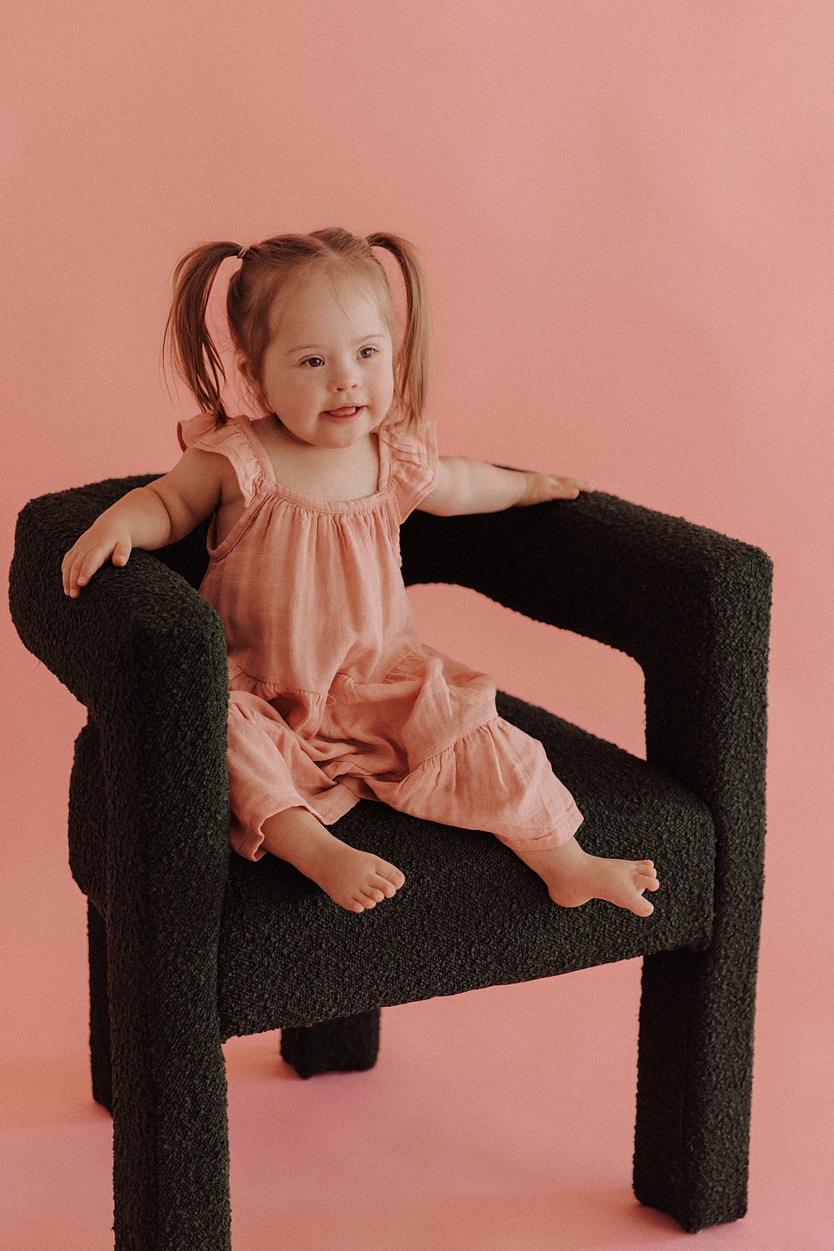 Toddler girl with blonde pigtails sits in a green chair in front of a pink backdrop
