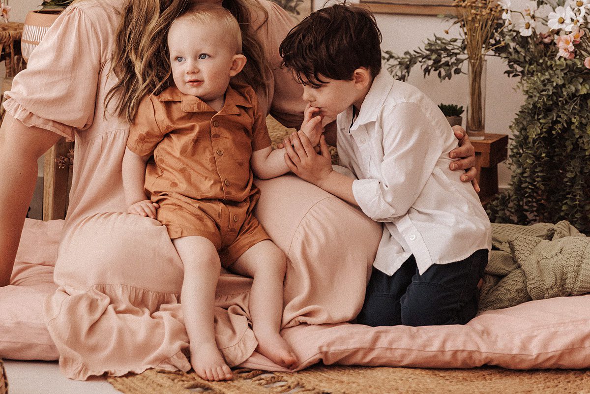 Baby boy sits in his mother’s lap while his older brother kisses his hand for a mini session picture
